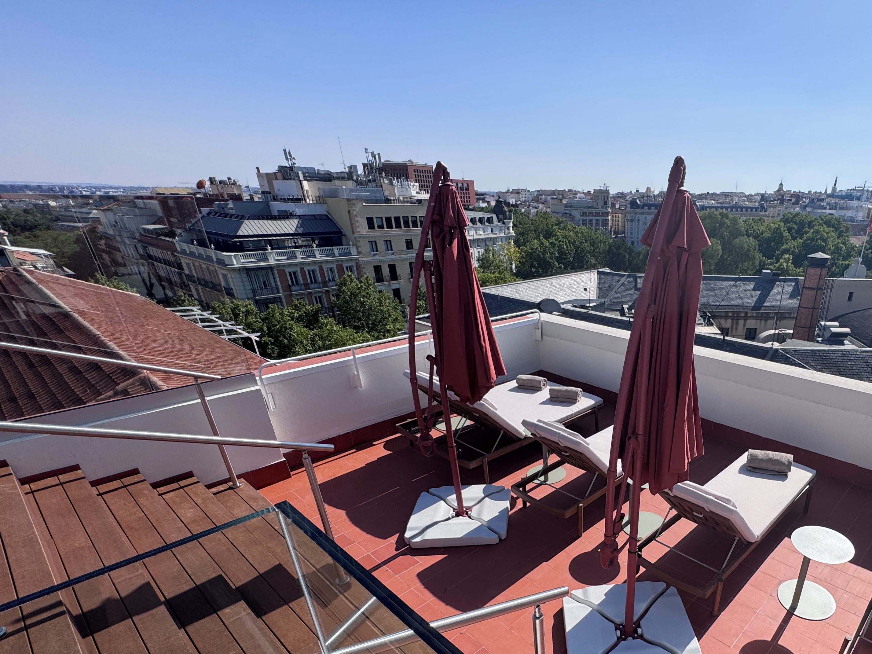Una terraza en la azotea con tumbonas, barandilla de cristal y vistas a la ciudad.