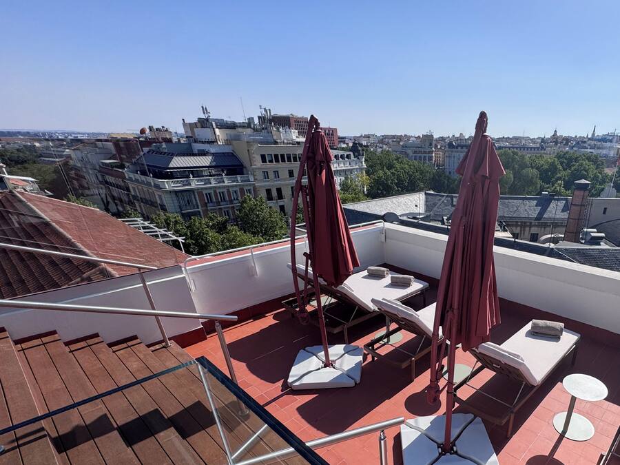 Una terraza en la azotea con tumbonas, barandilla de cristal y vistas a la ciudad.