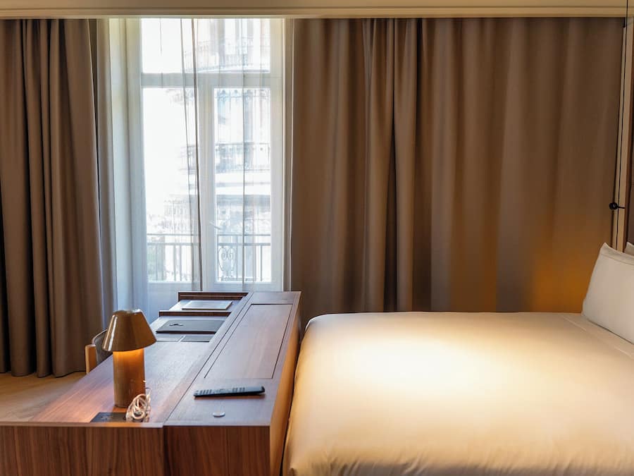Una habitación de hotel con cama, escritorio con lámpara y ventana con cortinas.