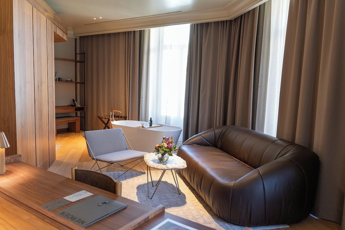 Een moderne woonkamer met een leren bank, een klein rond tafeltje met een bloemenvaas, een grijze fauteuil en een houten salontafel.