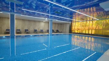 Una piscina interior con agua azul cristalina, sillas de descanso a un lado y un mural de un bosque en la pared.