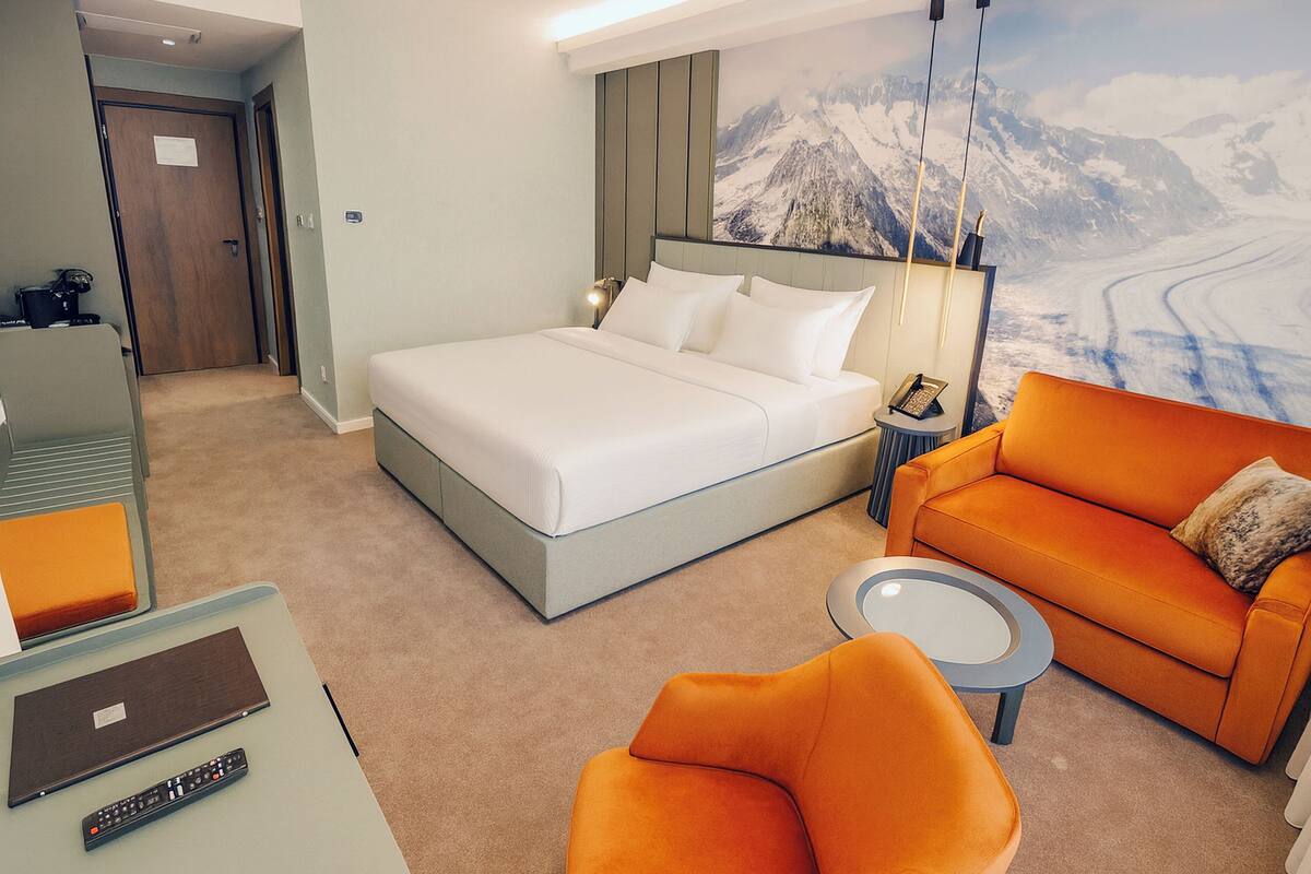 Habitación de hotel con cama, sofá naranja, una mesita redonda y un escritorio con computadora portátil y control remoto.