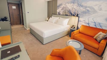 Ein Hotelzimmer mit einem Bett, einem orangefarbenen Sofa, einem kleinen runden Tisch und einem Schreibtisch mit Laptop und Fernbedienung.