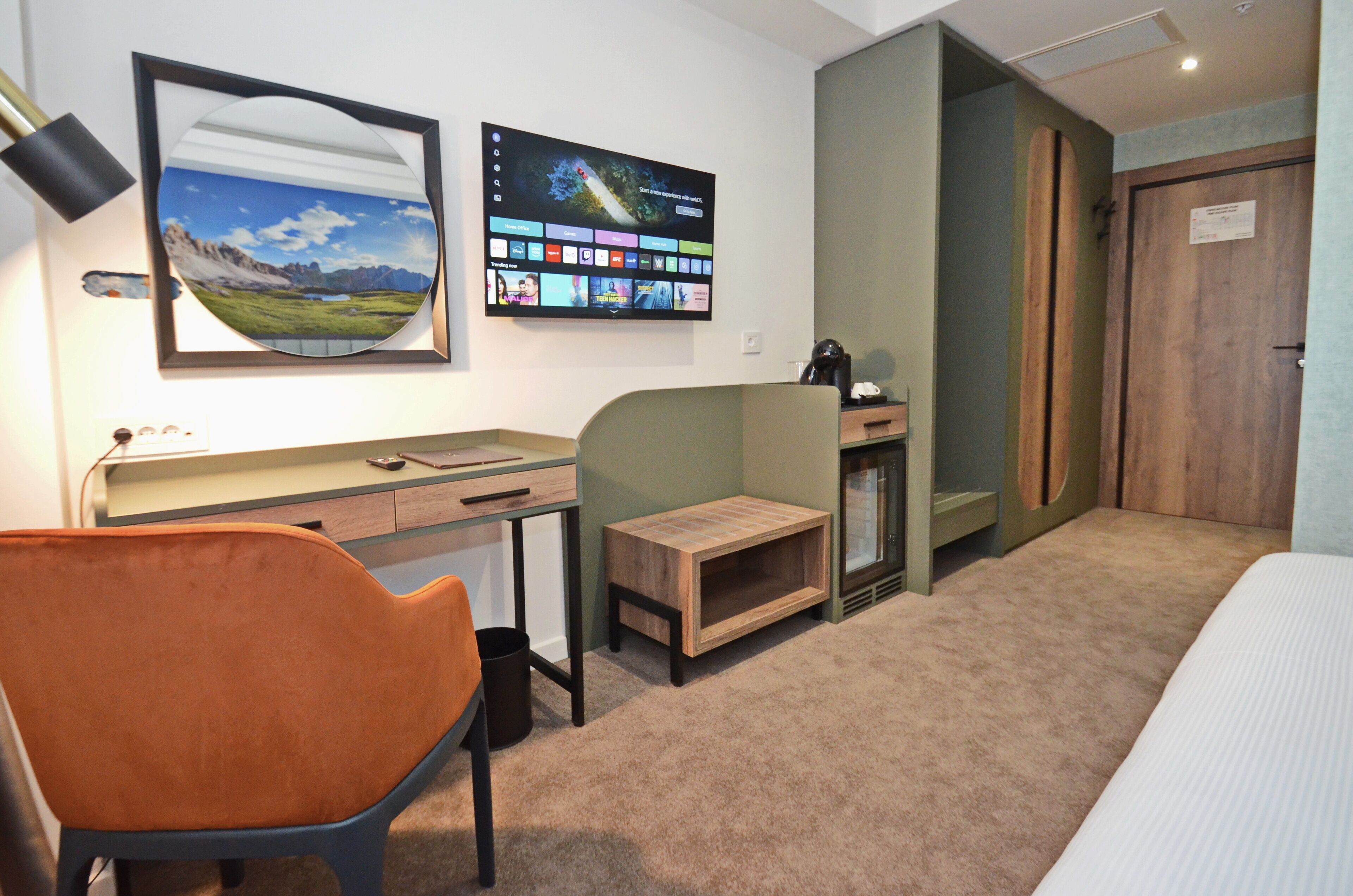Ein Hotelzimmer mit Schreibtisch, Stuhl, Bett und Fernseher, der ein Landschaftsbild sowie eine Speisekarte zeigt.