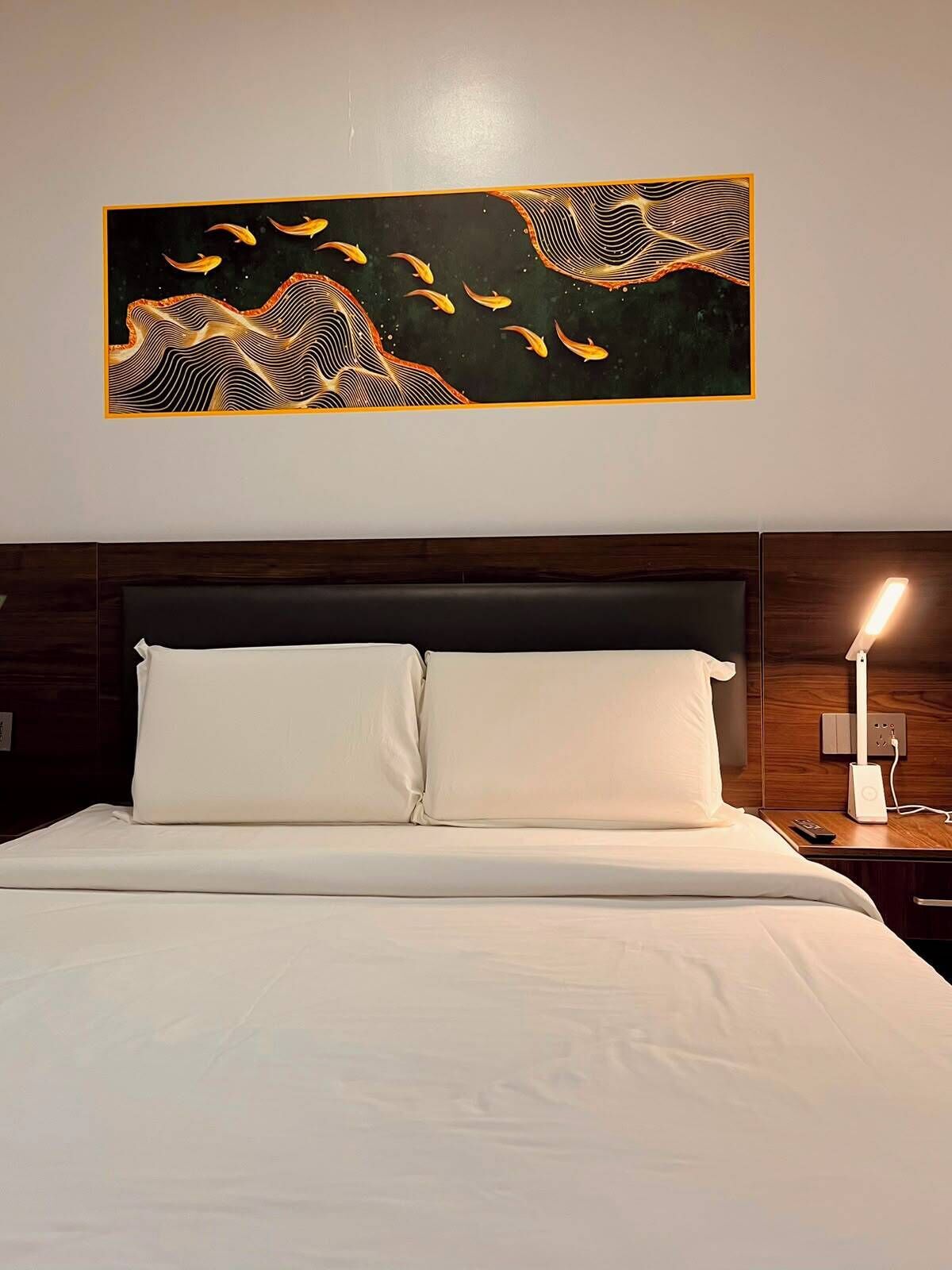 A hotel room with a bed, a nightstand with a lamp, and a wall-mounted artwork.