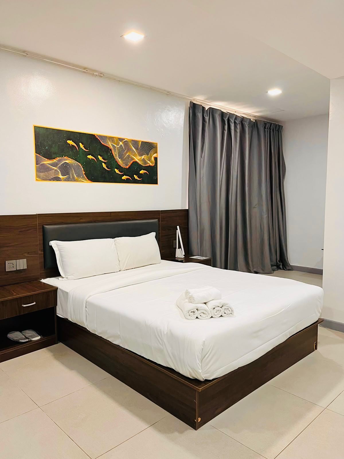 A hotel room with a large bed, a nightstand, a wall-mounted painting, and a window with curtains.