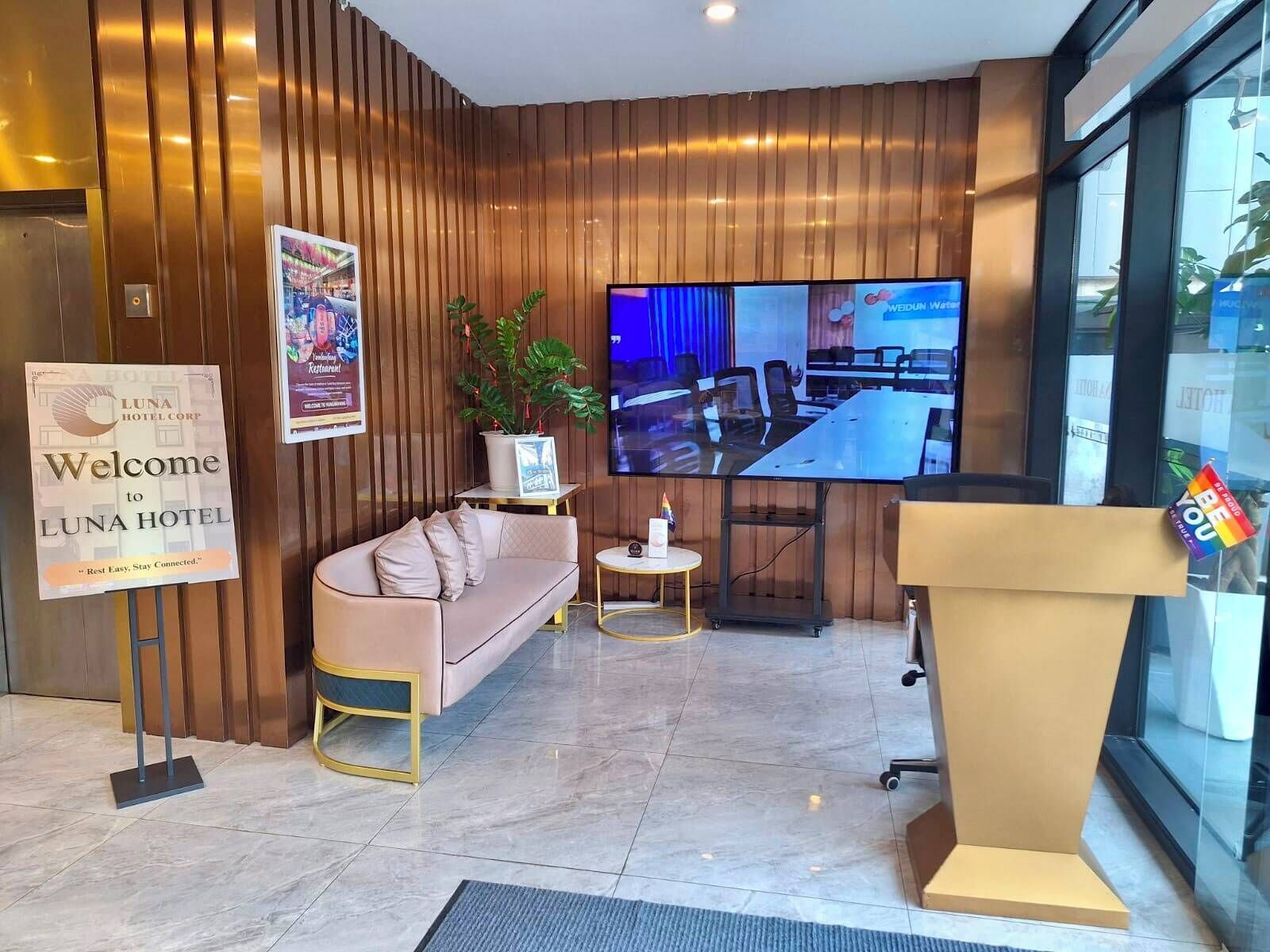 A hotel lobby with a reception desk, a sofa, a TV screen, and a sign welcoming guests to Luna Hotel.