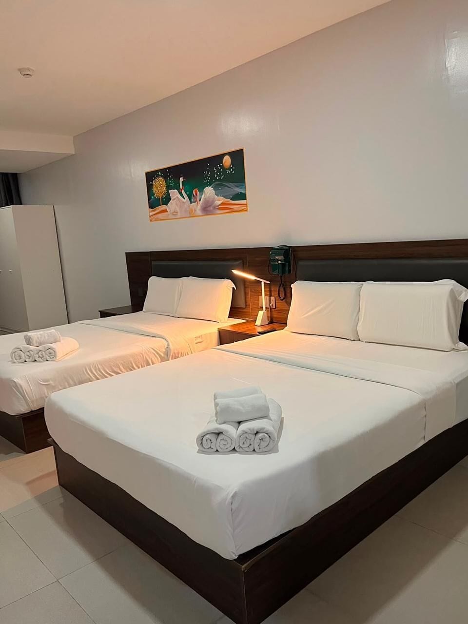 A hotel room with two beds, a painting on the wall, and a nightstand with a lamp.