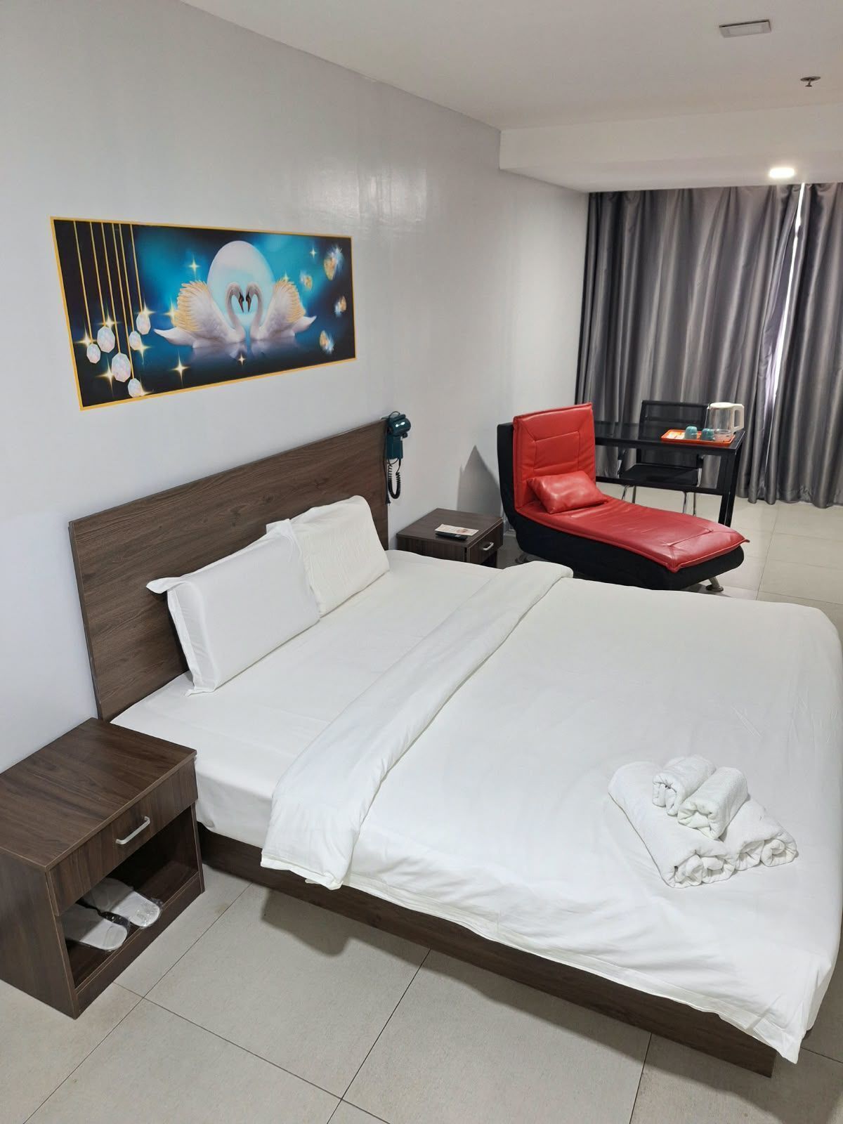 A hotel room with a large bed, a red chair, a small table with a kettle, and a painting on the wall.