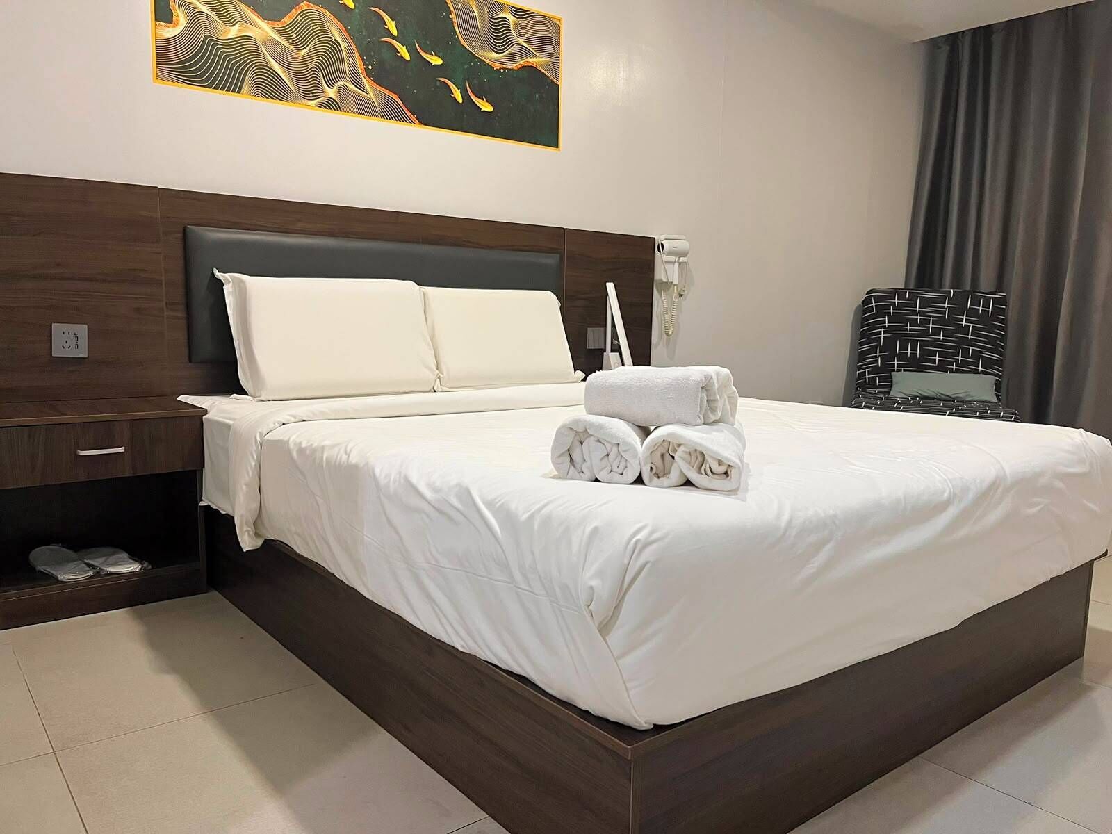 Ein Hotelzimmer mit einem großen Bett, einem Nachttisch, einem Hocker, einem fest montierten Telefon und einem Stapel Handtüchern.