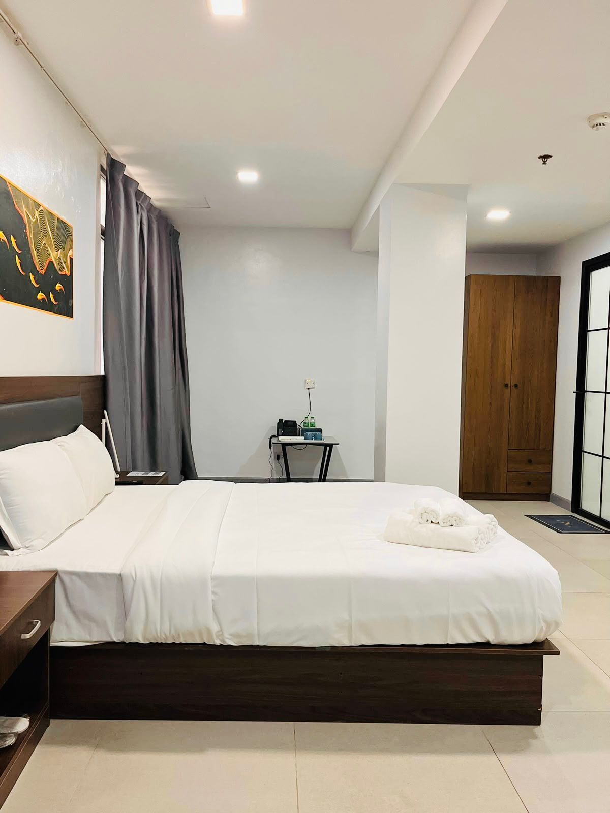 A modern hotel room with a large bed, a bedside table, a small desk with a chair, and a wardrobe.