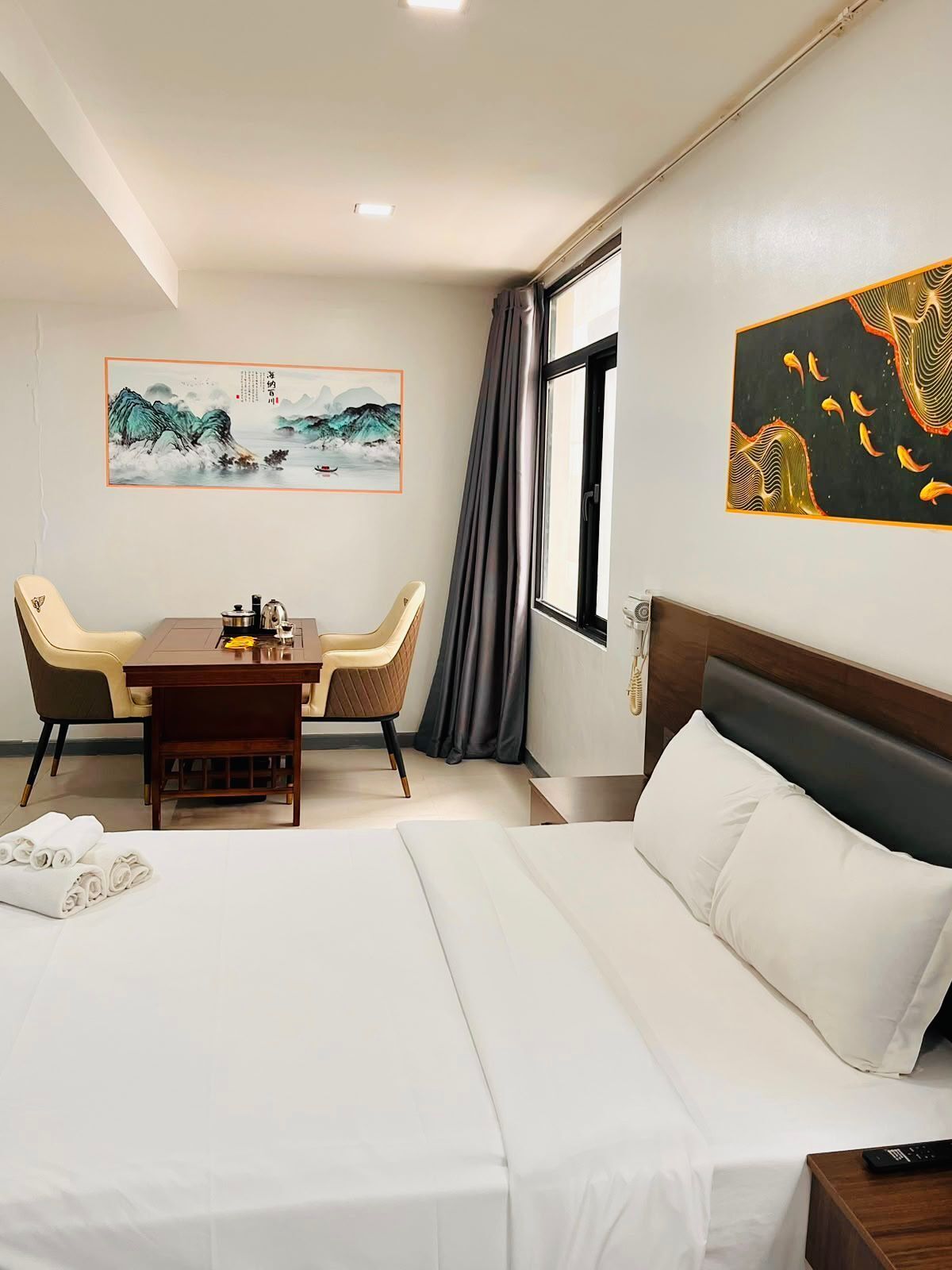 Une chambre d’hôtel avec un lit, un bureau avec des chaises, un tableau au mur et une fenêtre avec des rideaux.