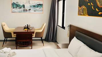 A hotel room with a bed, a desk with chairs, a painting on the wall, and a window with curtains.