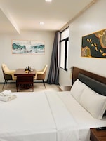 Une chambre d’hôtel avec un lit, un bureau avec des chaises, un tableau au mur et une fenêtre avec des rideaux.