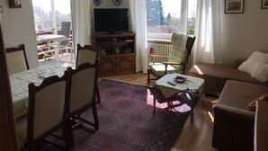 Una sala de estar con una mesa de comedor, sillas, un sofá, una televisión y vistas al exterior.
