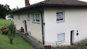 Una casa de un solo piso con tejado de tejas, una puerta principal y un pequeño jardín delantero con una silla y plantas en macetas.