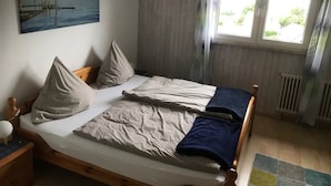 Une chambre à coucher avec un lit, des oreillers, une table de chevet, un tableau, une fenêtre avec des rideaux et un radiateur.