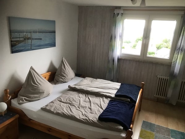 Une chambre à coucher avec un lit, des oreillers, une table de chevet, un tableau, une fenêtre avec des rideaux et un radiateur.