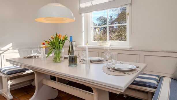 Comedor con una mesa blanca dispuesta para cuatro, un florero con flores y una ventana que da a vistas de árboles.