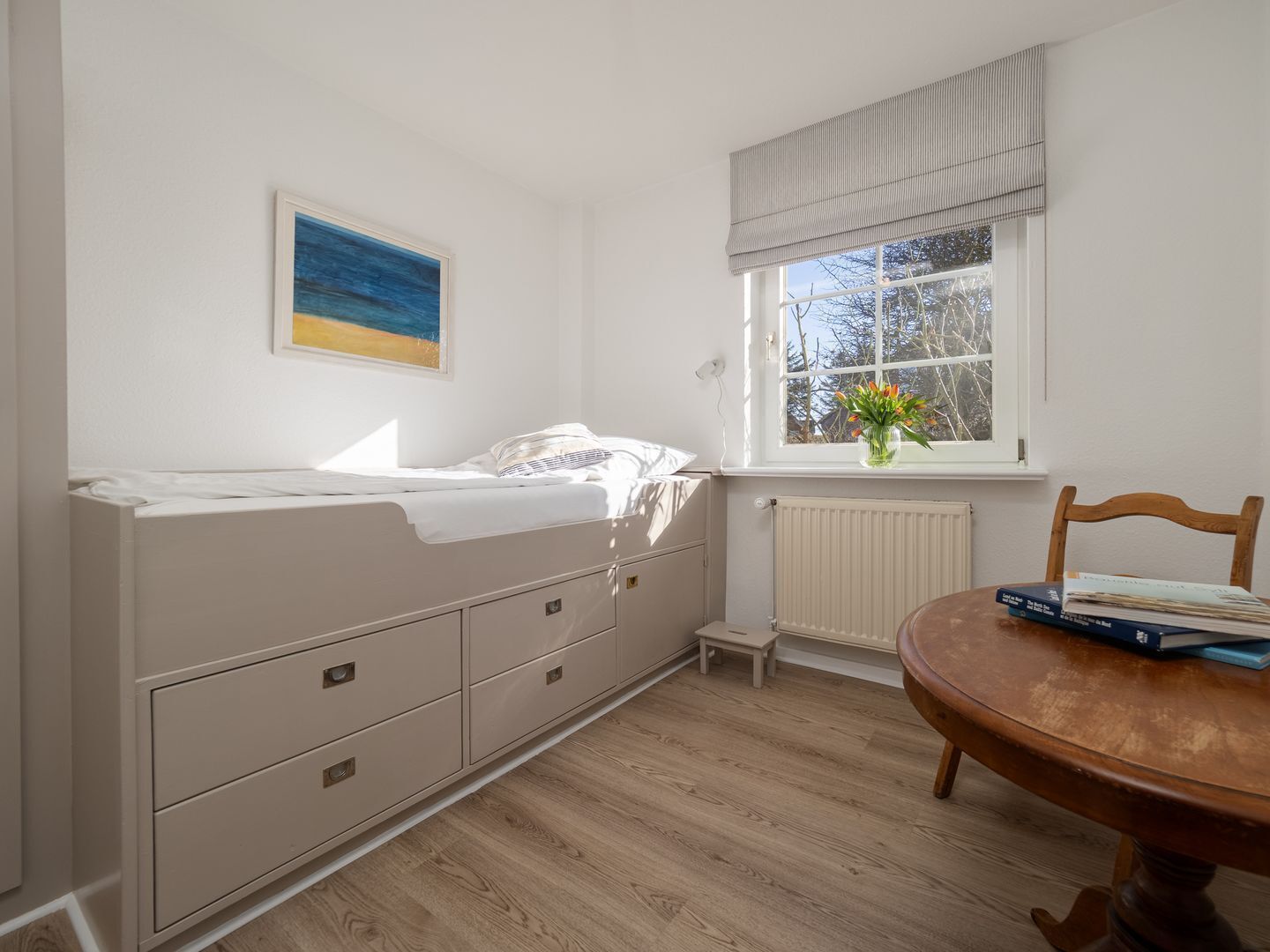 Ein Schlafzimmer mit einem Bett, einem kleinen Tisch mit Büchern, einem Fenster mit Aussicht und einer Heizung.