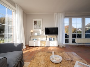 A living room with a sofa, a coffee table with a candle, a TV on a white cabinet, and large windows. - Amselhaus - Beautiful detached house with 120 m², 5 rooms and a large garden for up to (Sylt)