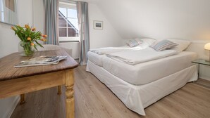 Un dormitorio con una cama, una mesita de noche con lámpara, una ventana con cortinas y un florero con flores.