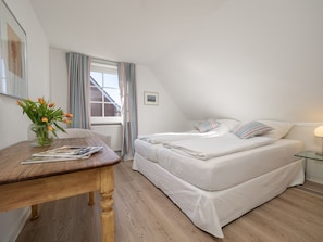 A bedroom with a bed, a bedside table with a lamp, a window with curtains, and a vase with flowers. - Amselhaus - Beautiful detached house with 120 m², 5 rooms and a large garden for up to (Sylt)