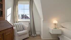Un dormitorio con un aparador de madera, un sillón a rayas, una mesita de noche con lámpara y una ventana con cortinas.