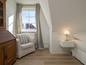 A bedroom with a wooden dresser, a striped armchair, a bedside table with a lamp, and a window with curtains. - Amselhaus - Beautiful detached house with 120 m², 5 rooms and a large garden for up to (Sylt)