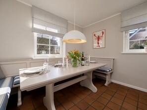 A dining area with a white table set for four, a bench, and a large window with curtains. - Amselhaus - Beautiful 120 m² single-family home with 5 rooms and a large yard, suitable for up to (Sylt)