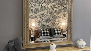 Une chambre avec un grand miroir, un papier peint à motifs floraux, un lit avec des coussins à carreaux, une petite figurine d’éléphant et un meuble décoratif avec une inscription.