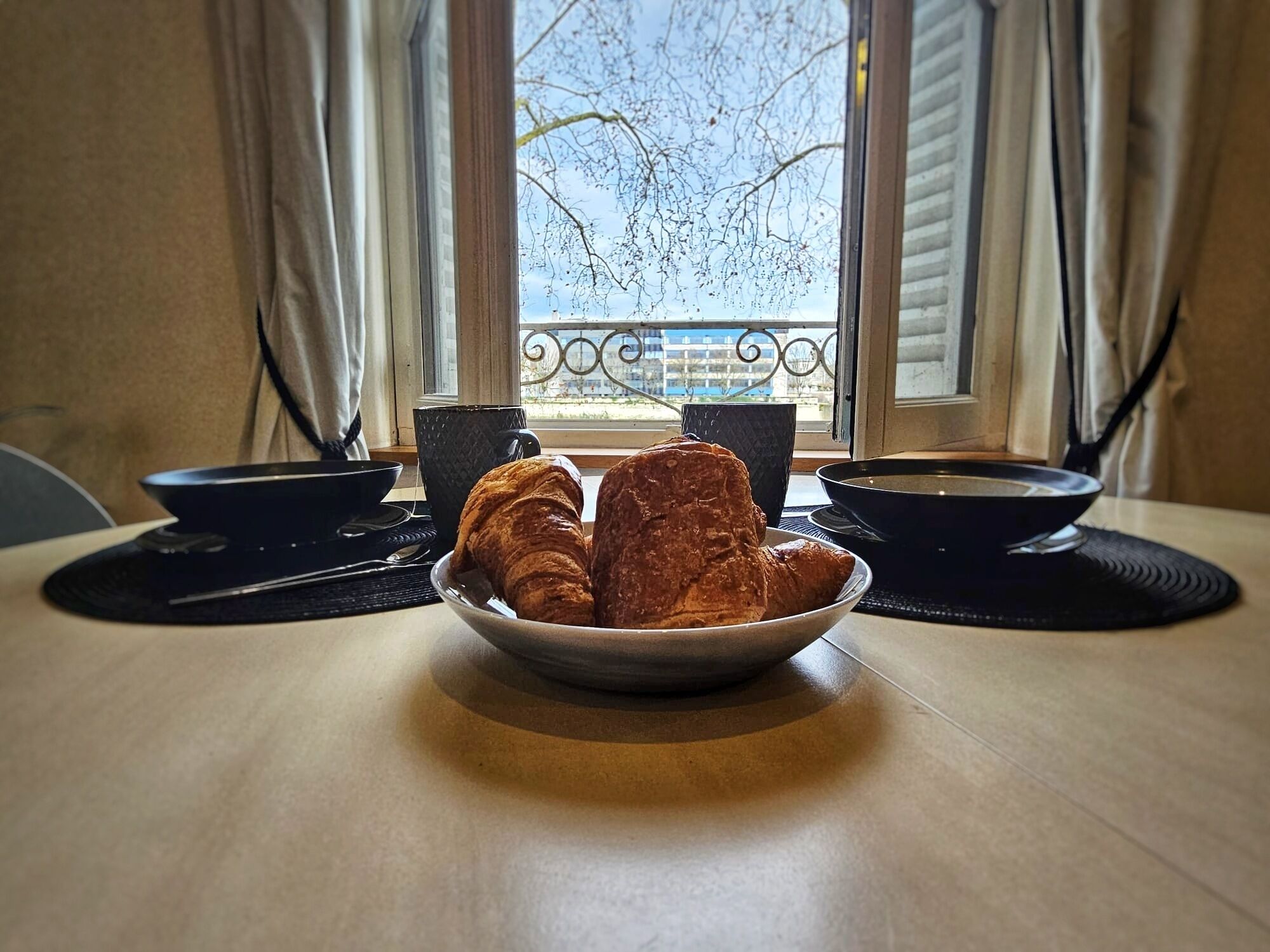 Una mesa con un bol de cruasanes, dos tazas negras y un bol de sopa.