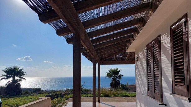 Un espacio al aire libre cubierto con vistas al mar, columnas de madera y una pared blanca con contraventanas.