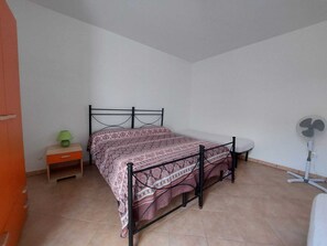 Una cama individual con colcha estampada en una habitación con suelo de baldosas, una mesita de noche con lámpara y un ventilador de pie.