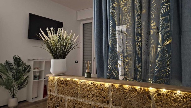 Un salon moderne avec un mur en pierre, une étagère blanche ornée d’une plante en pot, un téléviseur à écran plat et un rideau foncé à motifs de feuilles.