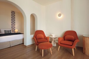 Una habitación de hotel moderna con una cama, dos sillones naranjas, una mesita redonda y una lámpara fijada a la pared.