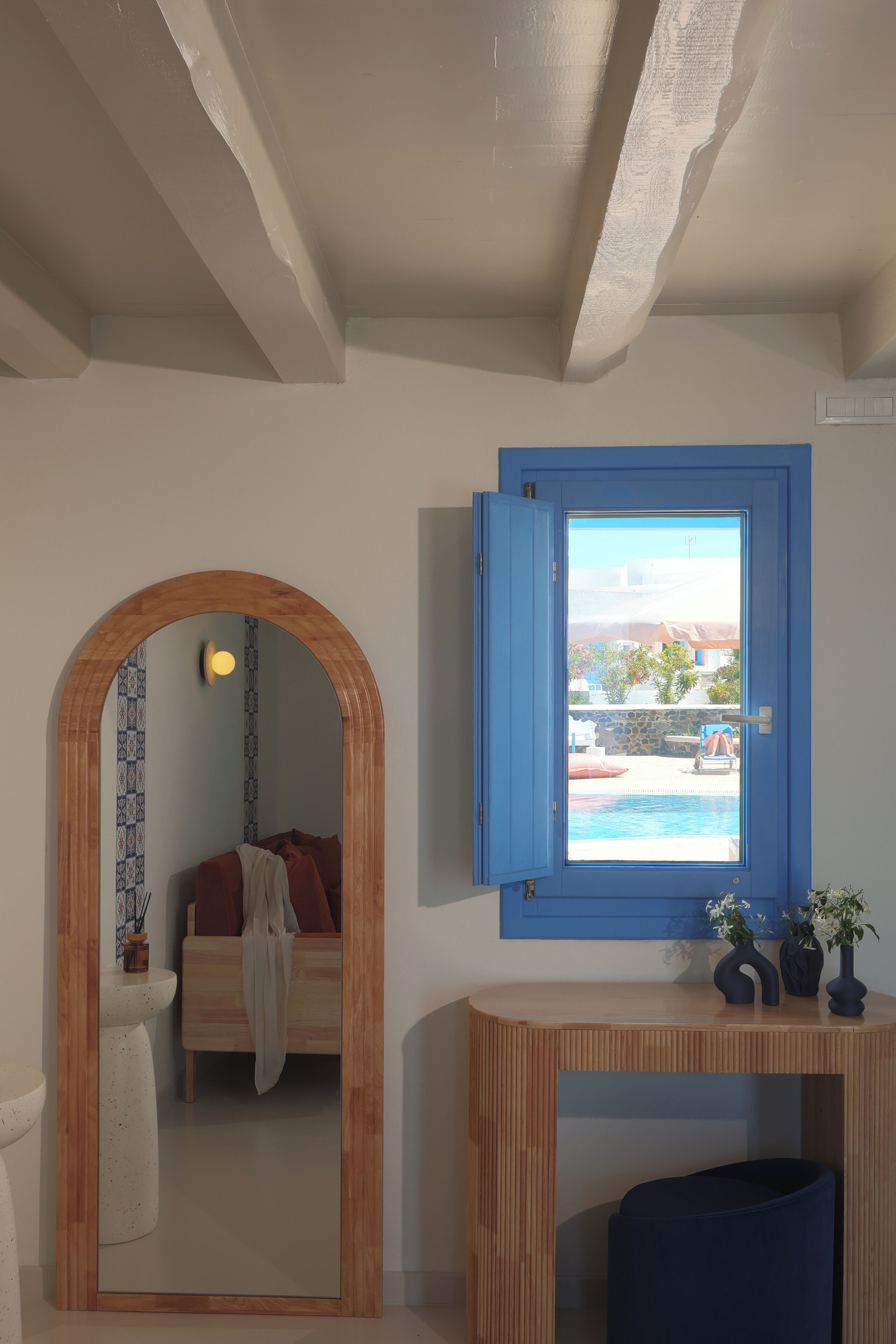 A room with a wooden ceiling, a blue-framed window, a wooden console table, and a bed with a red pillow.