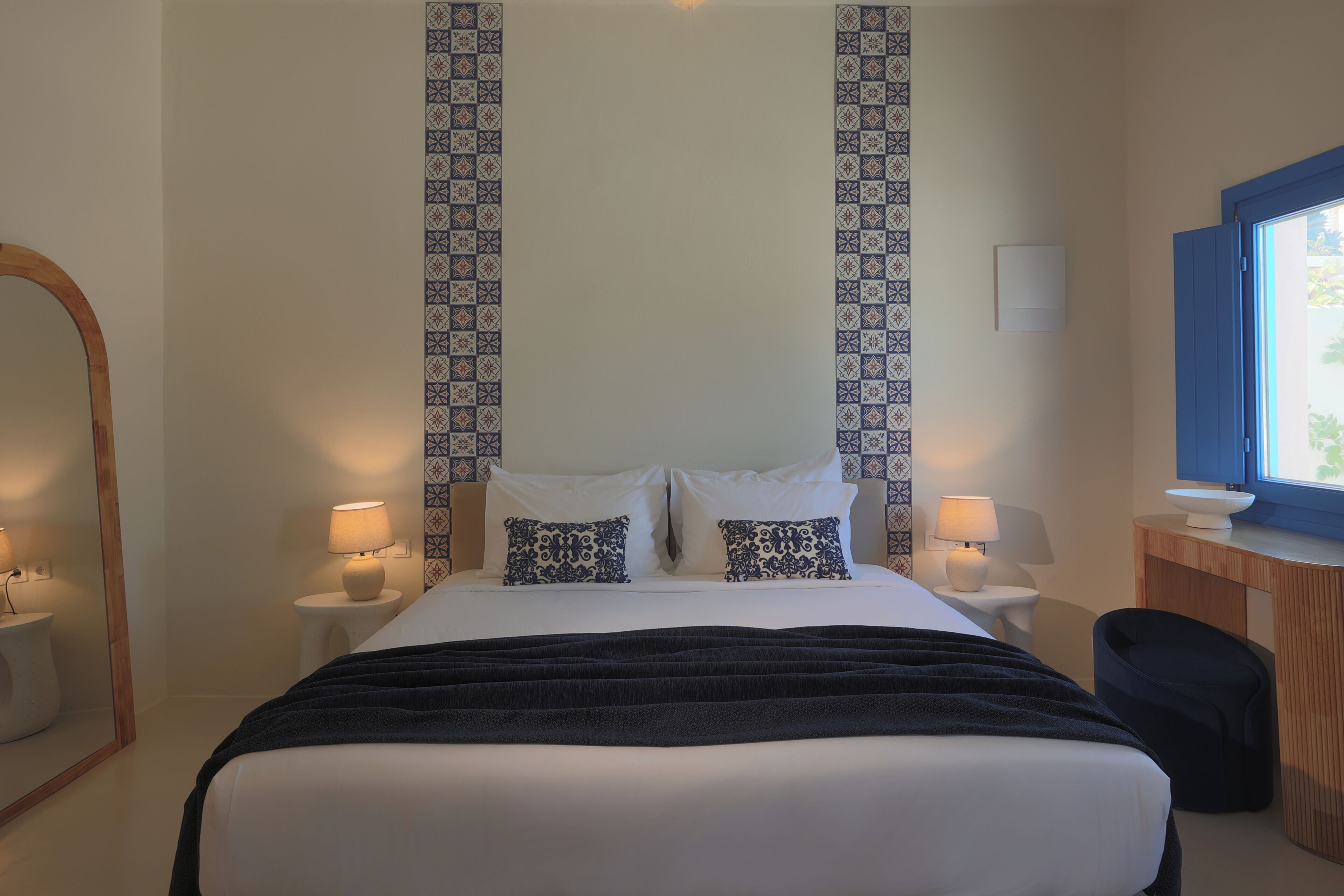 A hotel room with a bed, two bedside tables with lamps, a mirror, a dresser, and a window.