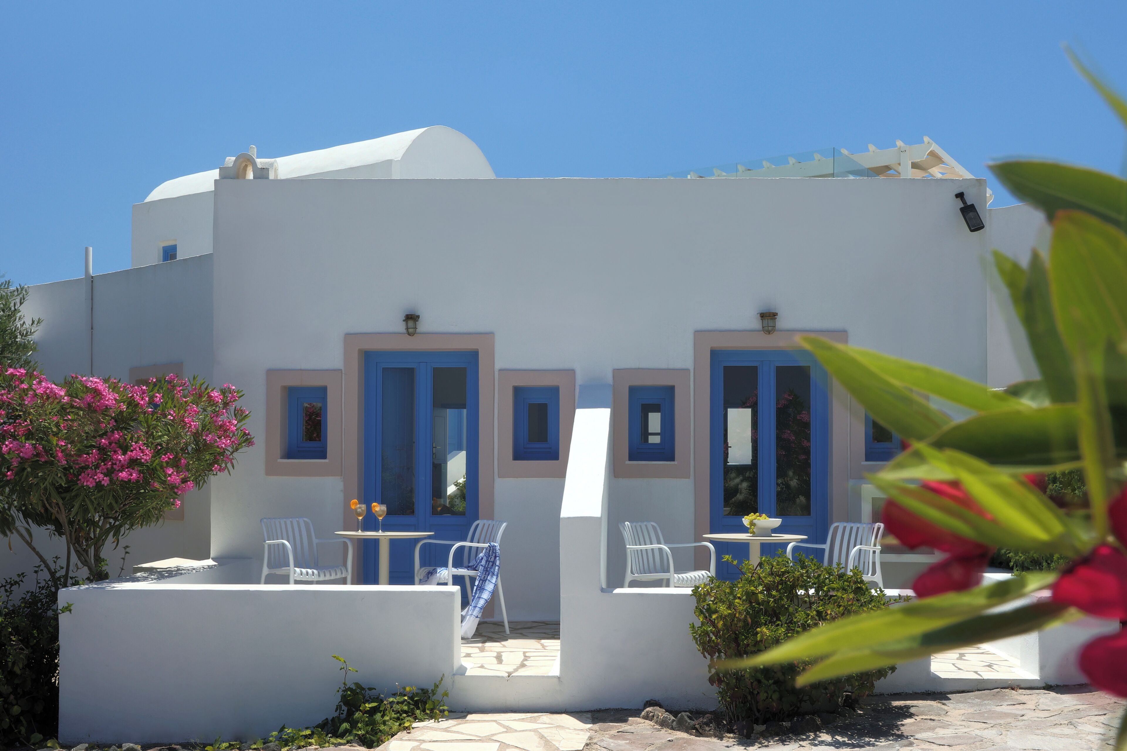 A white building with blue doors and windows, outdoor seating with white chairs, and a potted plant.