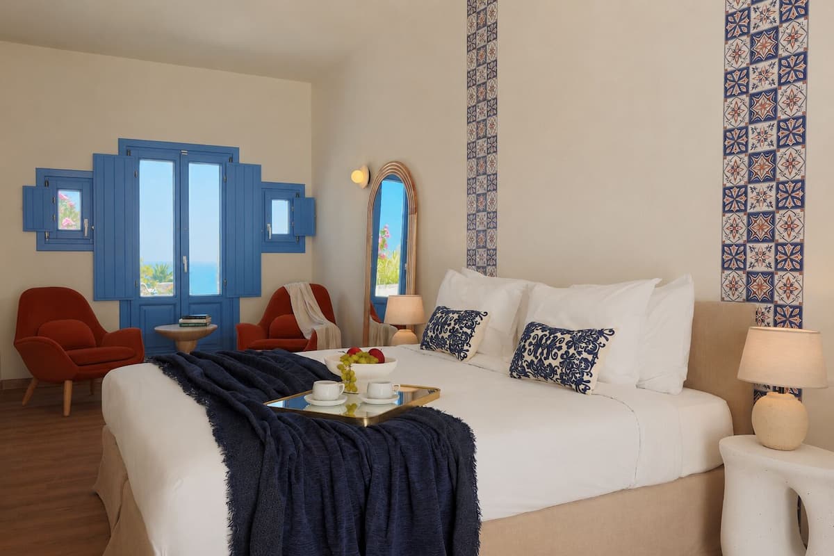 Een hotelkamer met een groot bed, twee stoelen, een kleine tafel en uitzicht op zee.