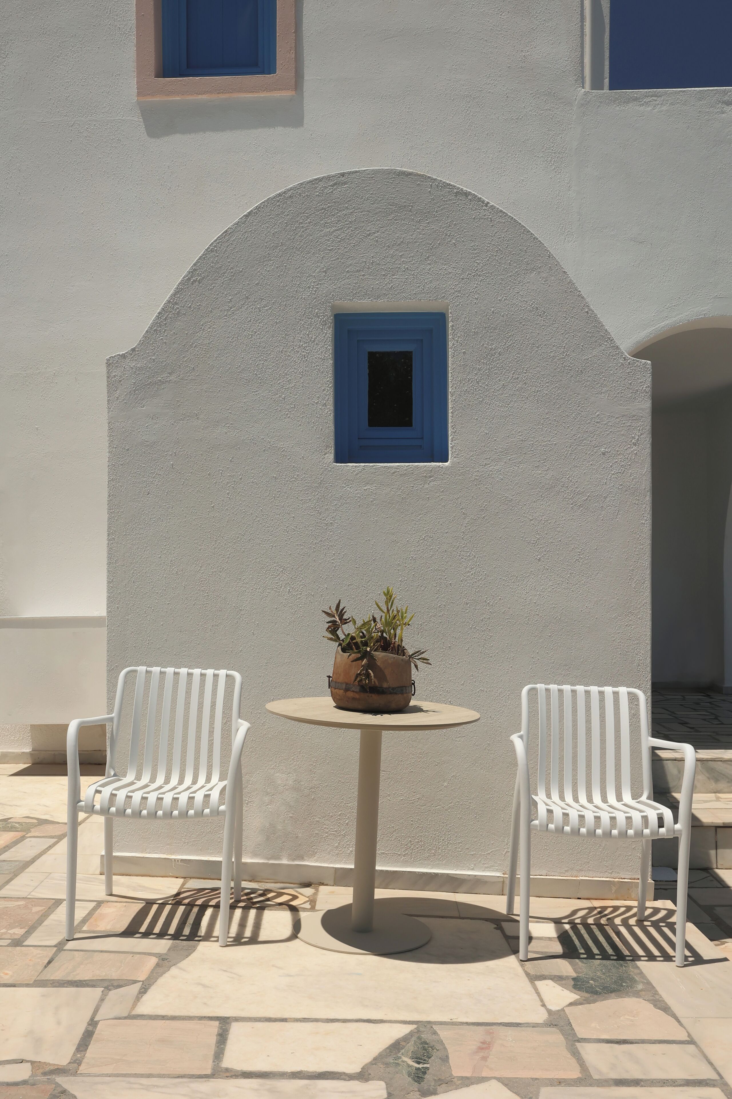 A white outdoor seating area with two chairs and a table, a potted plant, and a blue-framed window.