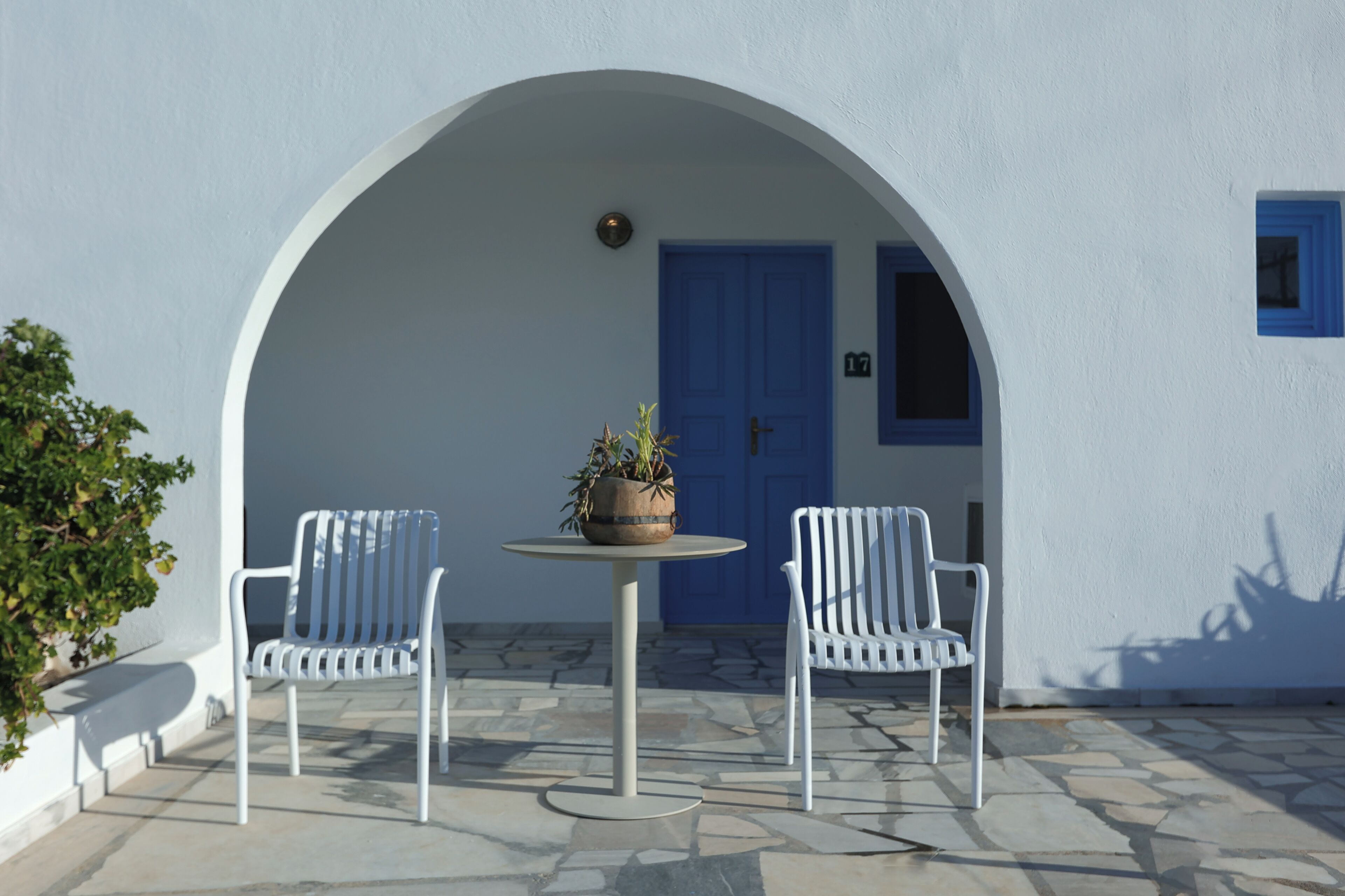 Un espacio exterior con dos sillas blancas y una mesa, una planta en maceta y una puerta azul.