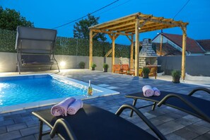 Ett poolområde med solstolar, en pergola och en eldstad i sten.