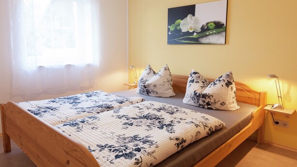 Une chambre à coucher avec un lit en bois, une literie à motifs floraux, une table de chevet avec une lampe et un tableau représentant une orchidée accroché au mur.