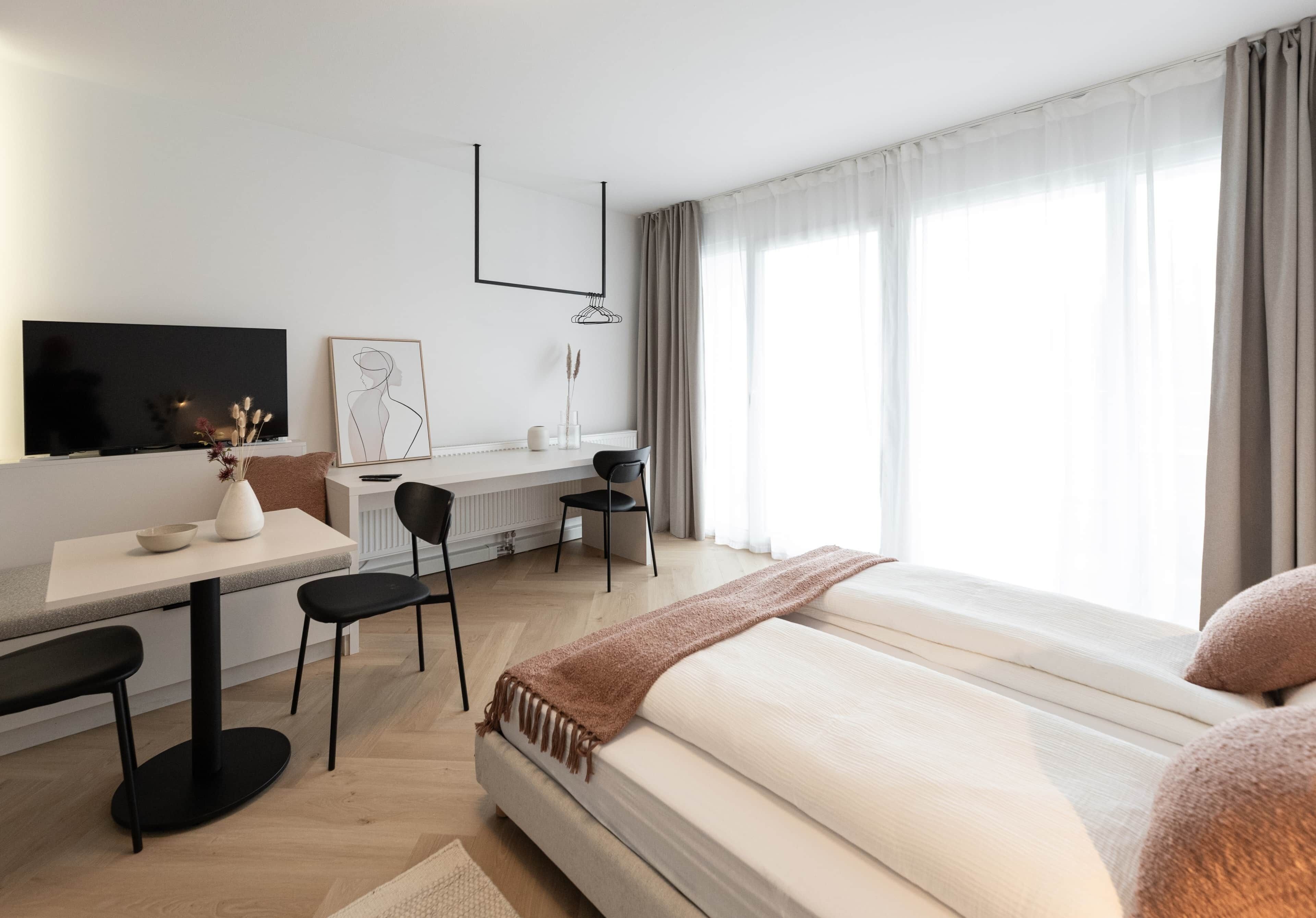 A modern hotel room with a bed, a desk, a chair, a TV, and a large window with curtains.
