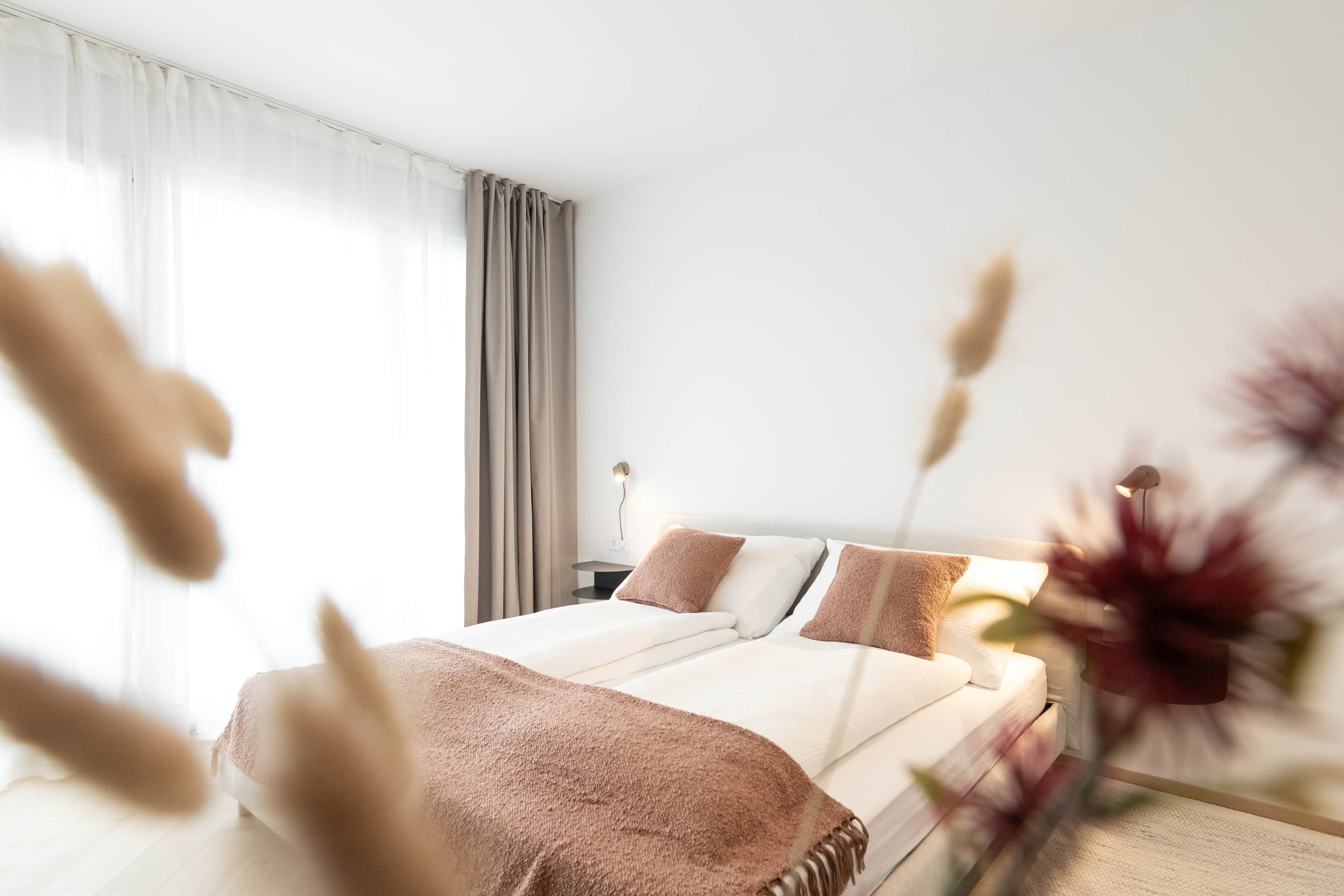 A modern hotel room with a large bed, a bedside table with a lamp, and a view of the outdoors through sheer curtains.