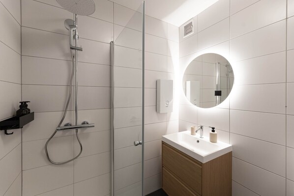 A modern bathroom with a glass-enclosed shower, a white sink with a wooden cabinet, and a round mirror with a light above it.