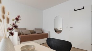 Ein schlicht eingerichtetes Schlafzimmer mit einem weißen Bett, einem runden Spiegel, einer weißen Kommode und einem schwarzen Stuhl.