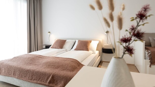 Ein modernes Hotelzimmer mit einem ordentlich bezogenen Bett, einem Nachttisch mit Lampe und einer Vase mit Trockenblumen.