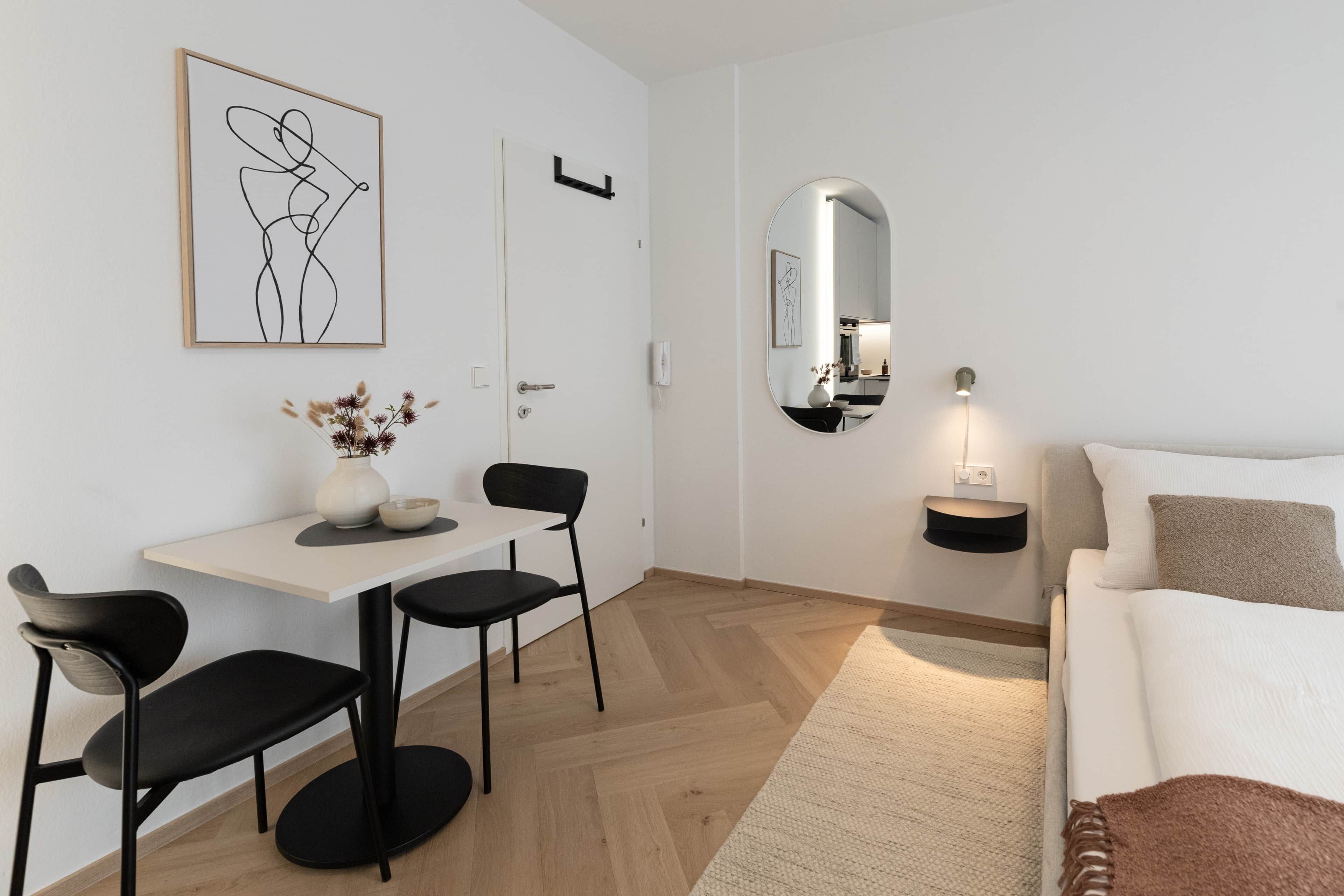 Una habitación moderna y minimalista con pared blanca, zona de comedor con mesa y sillas, sofá, espejo y una mesita auxiliar.